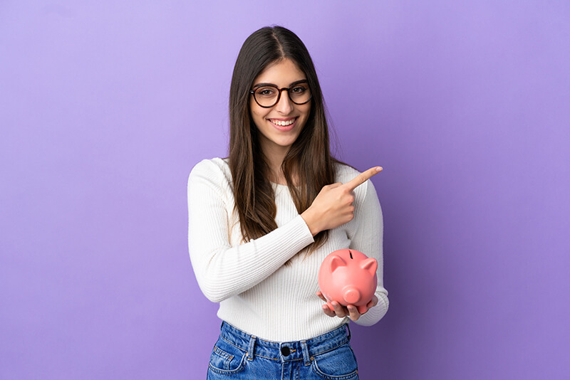 jovem mulher caucasiana segurando um cofrinho isolado em um fundo roxo apontando para o lado para apresentar um produto